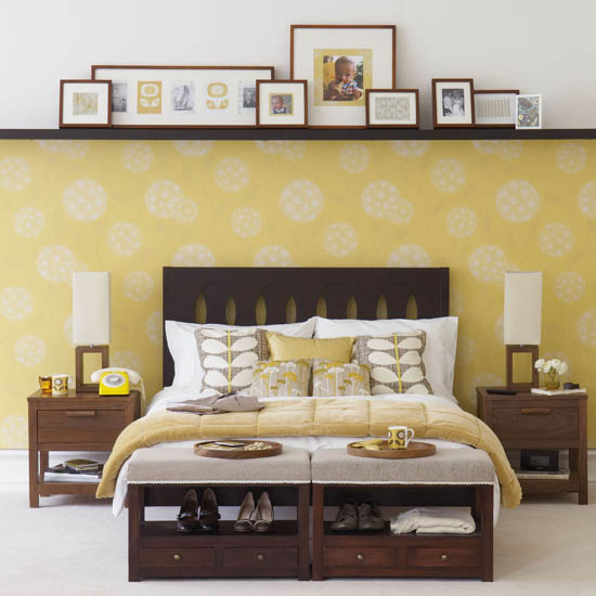 Yellow and white bedroom, floral wallpaper, shelf running along whole wall, framed pictures, double bed with dark wood headboard, two bedside tables, shoe storage at foot of bed, beige carpet. IH 07/2009 Pub orig