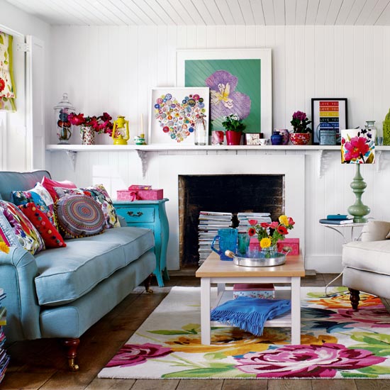 White living room, blue sofa with bright multicoloured cushions, bold floral rug, wooden coffee table, white sofa, stacked books used as table, stacked magazines in fireplace, wooden floorboards, 3D heart art on shelf, potted house plants. IH 06/2011 pub orig